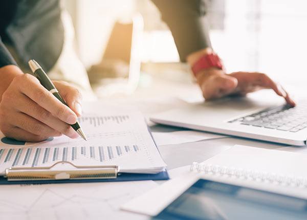 one hand holding a pen writing on graph printouts and the other using a laptop